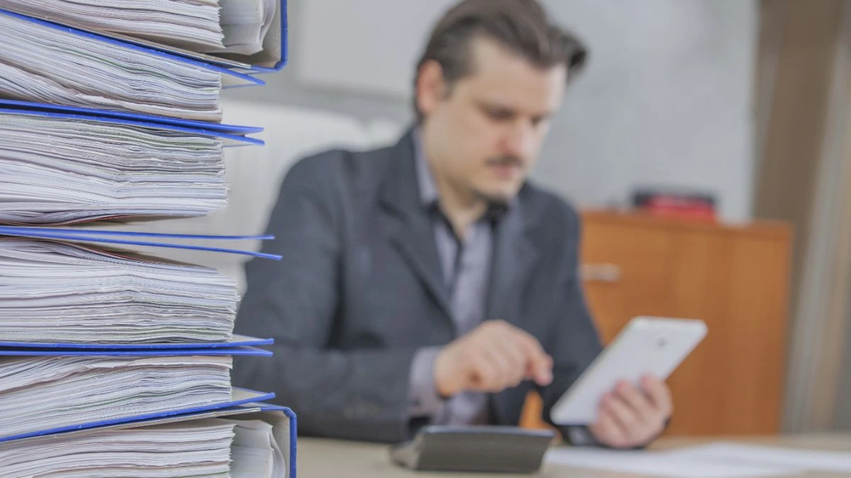 Como manter registros contábeis precisos e eficientes Homem sentado em uma mesa de escritório com pastas de documentos de resgistros contábeis ao lado, utilizando um tablet para trabalho. O ambiente é moderno e organizado.