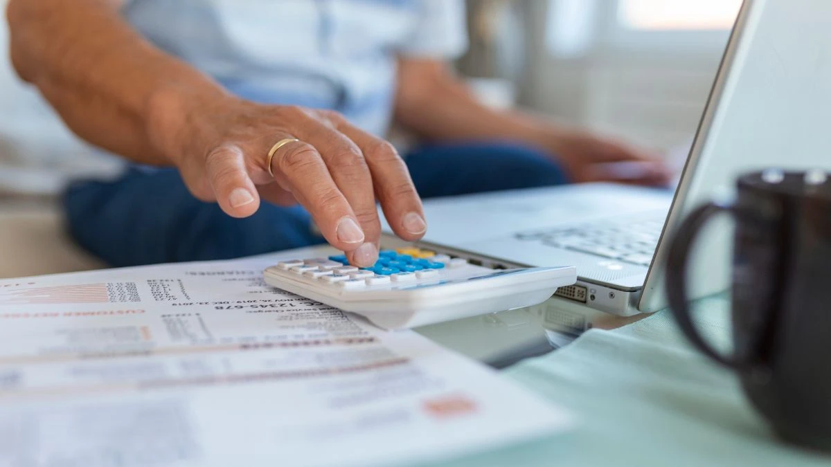Redução de custos com split de pagamento e automação Imagem mostrando uma pessoa calculando despesas em uma calculadora ao lado de um laptop e documentos em uma mesa. A imagem representa o gerenciamento financeiro e a organização de contas.