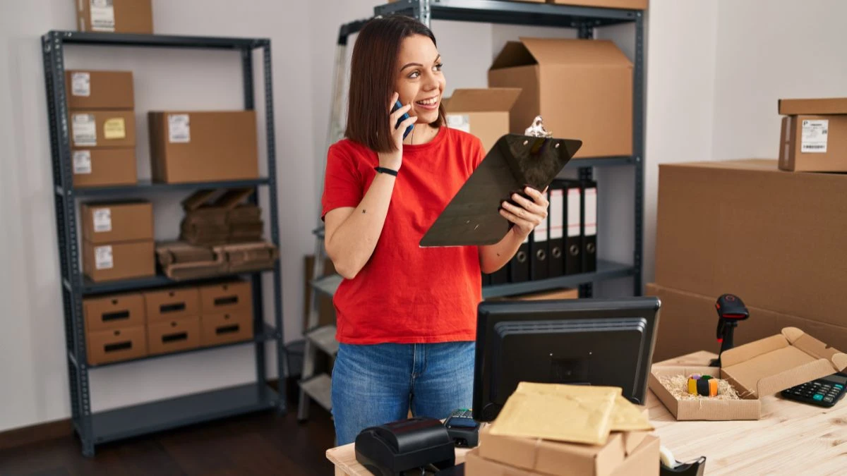 Controle de estoque: o que uma pequena empresa precisa saber Mulher sorridente em um ambiente de trabalho, usando um celular e segurando uma prancheta, com caixas e documentos ao fundo, representando logística e organização.