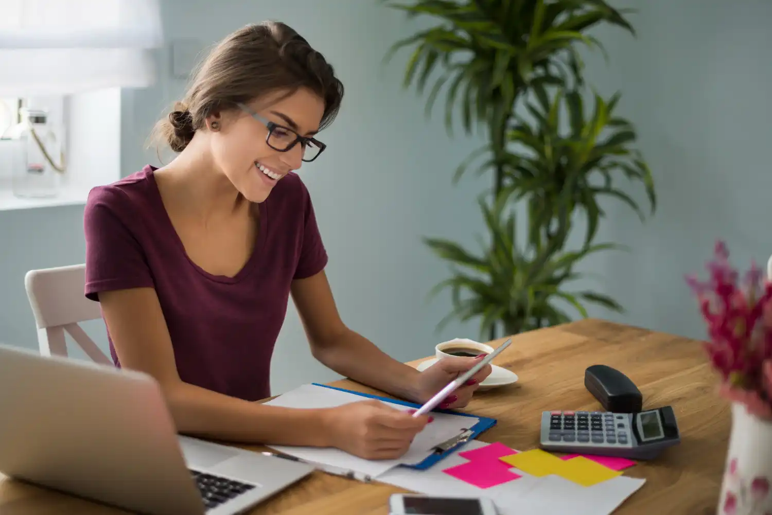 Mulher sorridente analisando opções de split de pagamento tributação em ambiente de escritório, com papéis, documentos, laptop, celular e calculadora na mesa.
